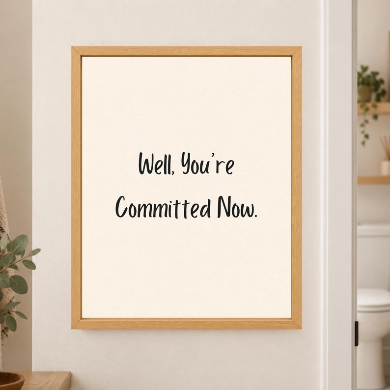 Framed minimalist bathroom wall art with the handwritten black phrase “Well, You’re Committed Now.” centered on a warm off-white background in a light wood frame beside a bathroom doorway.