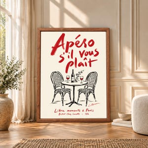 Framed French bistro wall art featuring red Apéro s'il vous plaît typography above two Paris cafe chairs and a small table with wine bottle, glasses and flowers on a warm cream background.