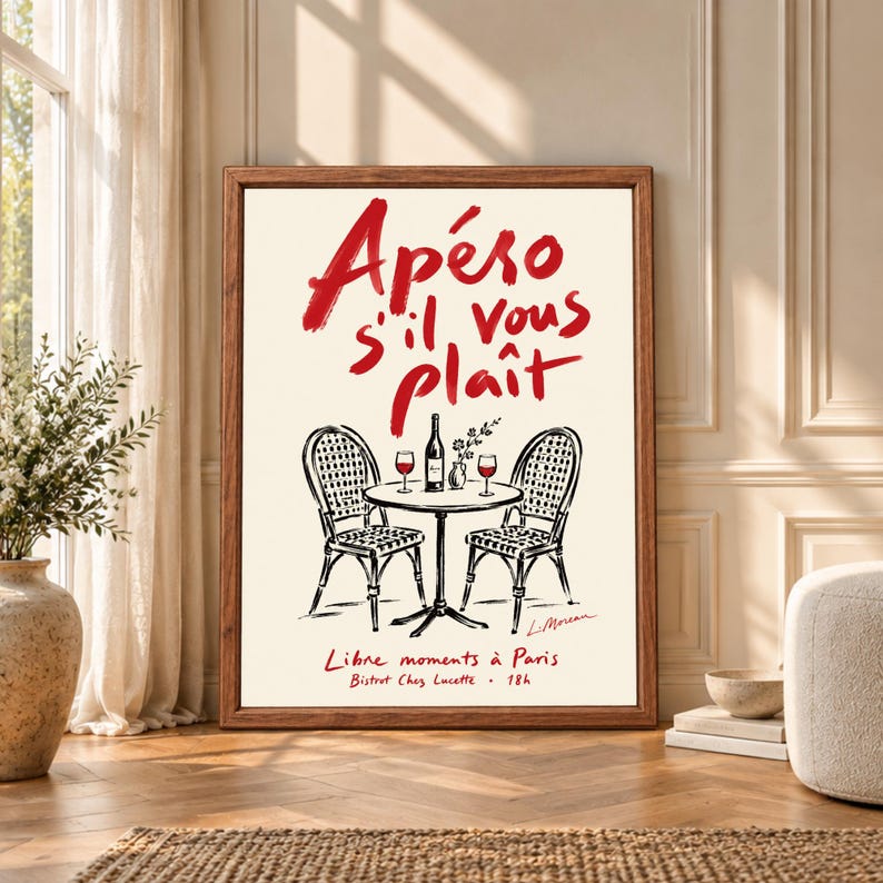 Framed French bistro wall art featuring red Apéro s'il vous plaît typography above two Paris cafe chairs and a small table with wine bottle, glasses and flowers on a warm cream background.