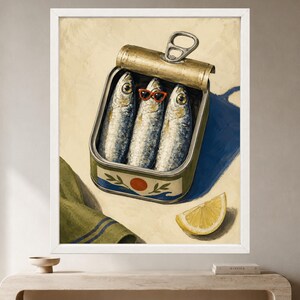 May include: A framed wall art print depicting an open metal tin containing three silver sardines. The center sardine wears small red-rimmed sunglasses. The tin features a red circle and green leaf graphic. A yellow lemon wedge sits nearby.