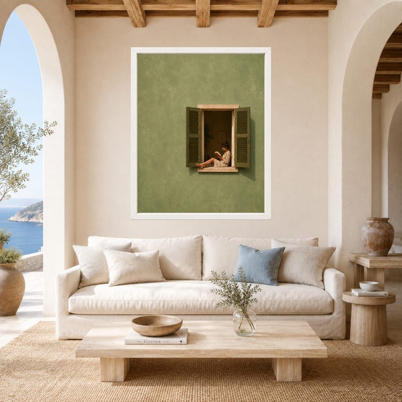 May include: A framed wall art print featuring a sage green exterior wall with an open window. A person sits on the window ledge reading a book. The artwork is displayed in a white frame, centered above a beige sofa with decorative pillows.