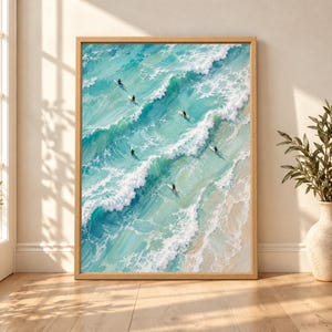 Framed aerial surfers wall art showing six tiny surfers paddling through textured turquoise ocean waves with white foam rolling toward a pale sandy beach.