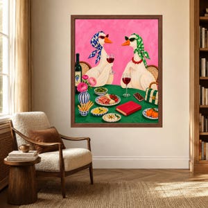 May include: A framed wall art print featuring two white geese wearing headscarves and sunglasses, sitting at a green table with wine, appetisers, and a red book. The background is a solid pink, and the geese are positioned as if dining together.