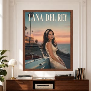 Framed Lana Del Rey wall art showing a female singer in a cream satin dress leaning against a turquoise vintage convertible on a California coastal road at sunset with the ocean behind her.