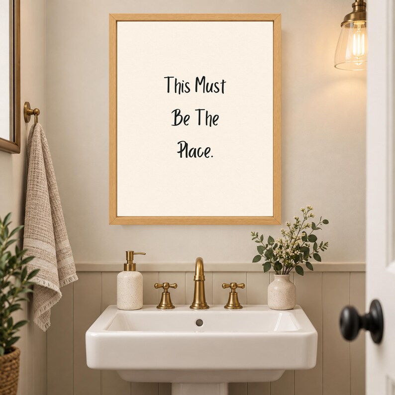 May include: A rectangular wall art print in a light wood frame featuring the black text This Must Be The Place centered on a cream background. The print is displayed above a white bathroom sink with gold fixtures and a beige towel hanging nearby.
