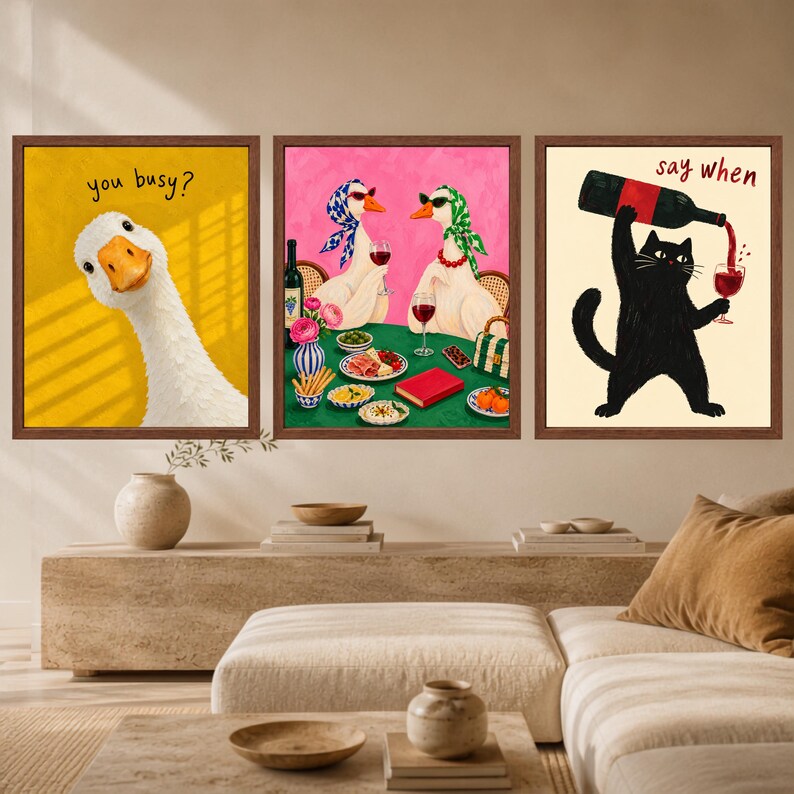 May include: A set of three framed wall art prints. The left print shows a white goose on a yellow background with the text 'you busy?'. The centre print depicts two geese wearing headscarves at a table with wine and food. The right print shows a black cat pouring red wine into a glass with the text 'say when'.
