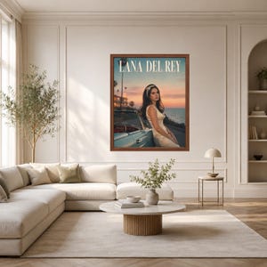 May include: A framed wall art print featuring a portrait of Lana Del Rey leaning against a light blue vintage convertible car at sunset by the ocean. The text LANA DEL REY is printed in white at the top of the image, which is displayed on a wall.