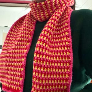 May include: A hand-crocheted scarf featuring a repeating pattern of vibrant pink, bright yellow, and deep red yarn. The scarf has a textured, woven appearance with a solid pink border along the edges and is draped around the neck.