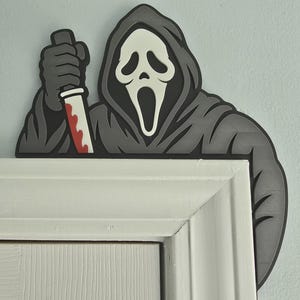 May include: A flat, decorative door topper featuring a stylized figure in a dark grey hooded robe with a white ghost-like mask. The figure holds a knife with a silver blade and red blood-like markings, designed to sit atop a door frame corner.