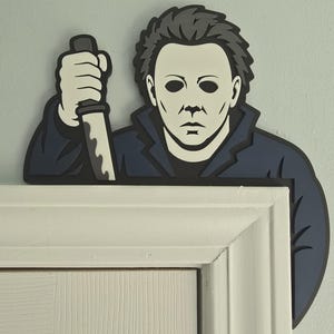 Puede incluir: Un adorno de pared plano diseñado para colocarse sobre el marco de una puerta, con una ilustración estilizada de un personaje con máscara blanca, cabello oscuro y chaqueta azul oscuro, sosteniendo un cuchillo plateado en la mano alzada.