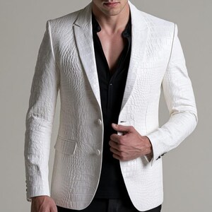 Può includere: Un blazer bianco con una texture in rilievo effetto pelle di coccodrillo, revers classici e due bottoni frontali. Il blazer è indossato sopra una camicia nera. Il capo presenta maniche lunghe con polsini abbottonati e due tasche con patta.