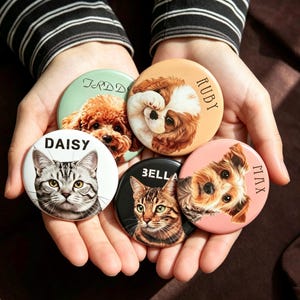 May include: Five circular pin-back buttons featuring illustrations of pets. The buttons display portraits of a poodle, a spaniel, a tabby cat, a Bengal cat, and a terrier, with the names Teddy, Ruby, Daisy, Bella, and Max printed on them.