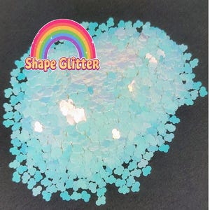 Puede incluir: Primer plano de purpurina azul iridiscente con forma de nubes. La purpurina está esparcida sobre una superficie negra. El texto "Shape Glitter" es visible en la imagen.