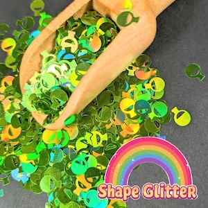 Puede incluir: Purpurina verde con forma de alienígena que se derrama de una cuchara de madera. La purpurina tiene un brillo holográfico, que refleja varios colores. Una pegatina de arcoíris con las palabras "Shape Glitter" está en la esquina inferior derecha.