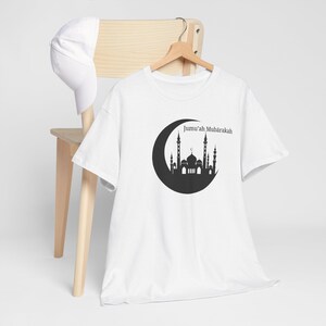 Op de afbeelding: Een wit T-shirt met korte mouwen en een zwarte afbeelding van een halve maan met daarin het silhouet van een moskee en de tekst Jumu'ah Mubārakah. Een effen witte baseballpet ligt op de houten rugleuning van de stoel achter het T-shirt.