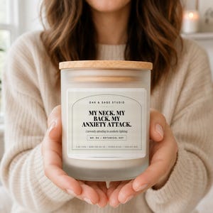 May include: A frosted glass candle jar with a light wood lid and a white label featuring the text: OAK & SAGE STUDIO, MY NECK. MY BACK. MY ANXIETY ATTACK. Currently spiraling in aesthetic lighting. NO. 04 - BOTANICAL SOY. 11 OZ / 311G - BURN TIME 50+ HR.