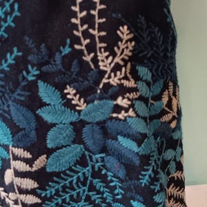 May include: A dark navy blue skirt with embroidered botanical patterns. The design features teal and light beige leaves and branches. The skirt's hem is adorned with a detailed floral pattern, creating a textured and visually appealing garment.