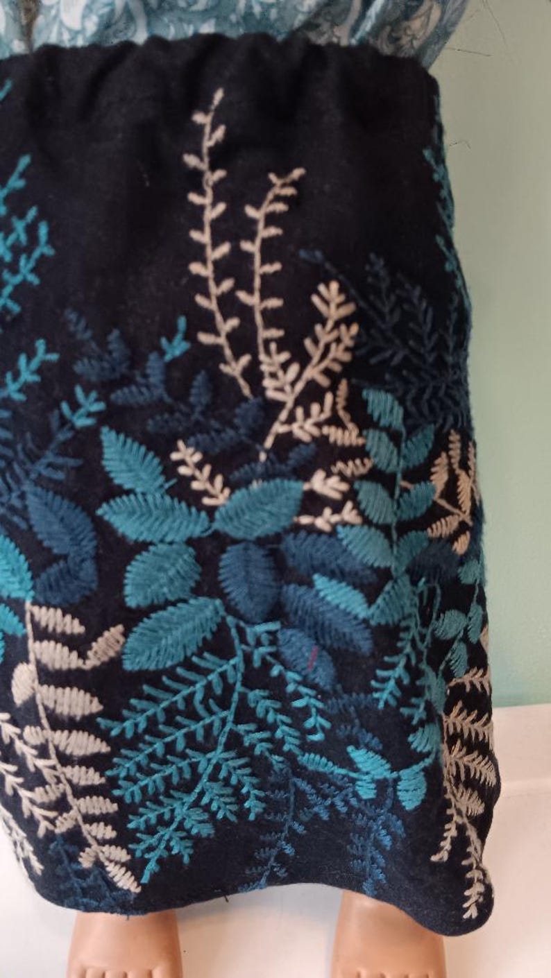May include: A dark navy blue skirt with embroidered botanical patterns. The design features teal and light beige leaves and branches. The skirt's hem is adorned with a detailed floral pattern, creating a textured and visually appealing garment.