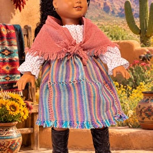 May include: A doll with dark braided hair wearing a white long-sleeved shirt with a small floral pattern, a pink fringed shawl, a multi-colored striped skirt with a fringed hem, and black boots. The doll has brown skin and small hoop earrings.