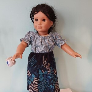 May include: A doll wearing a blue floral print top and a black skirt with teal and beige leaf embroidery. The doll holds a small teacup with a blue floral design. The doll has dark hair and is standing on a white surface.