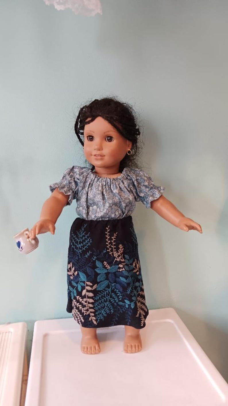 May include: A doll wearing a blue floral print top and a black skirt with teal and beige leaf embroidery. The doll holds a small teacup with a blue floral design. The doll has dark hair and is standing on a white surface.