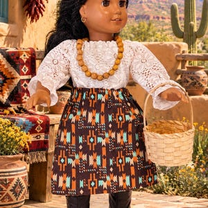 May include: A doll with dark braided hair wearing a white lace long-sleeve top, a brown skirt with a geometric pattern in orange and turquoise, a wooden bead necklace, and black boots. The doll holds a small, round, light-colored woven basket.