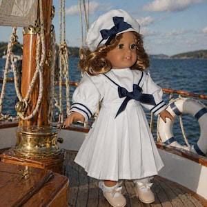 May include: A doll dressed in a white sailor outfit with navy blue accents, including a hat and bow. The doll is standing on a wooden boat deck with a blue ocean and sky in the background. The doll is wearing beige shoes.