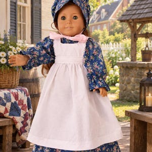 May include: A doll with brown hair and brown eyes wearing a blue floral print dress with long sleeves, a white apron, and a matching blue floral bonnet tied with a light pink ribbon under the chin. The doll has a light skin tone and a neutral expression.