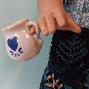 May include: A small, off-white ceramic creamer with a blue heart design and a handle. The creamer is held by a doll wearing a blue floral top and a black skirt with blue and beige leaf embroidery.