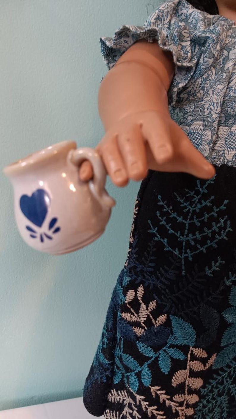 May include: A small, off-white ceramic creamer with a blue heart design and a handle. The creamer is held by a doll wearing a blue floral top and a black skirt with blue and beige leaf embroidery.