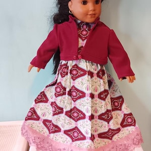 May include: A doll wearing a burgundy bolero jacket over a patterned dress. The dress features a white background with a repeating burgundy and pink floral design. The dress has a pink lace trim.