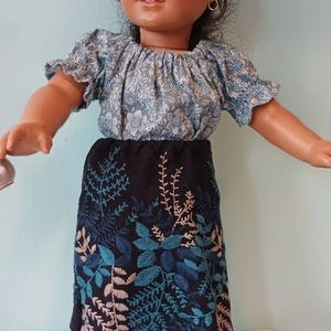 May include: A doll wearing a blue floral top and a black skirt with blue and white embroidered leaf designs. The doll has dark hair and brown eyes, and is wearing gold hoop earrings. The doll is standing on a white surface.