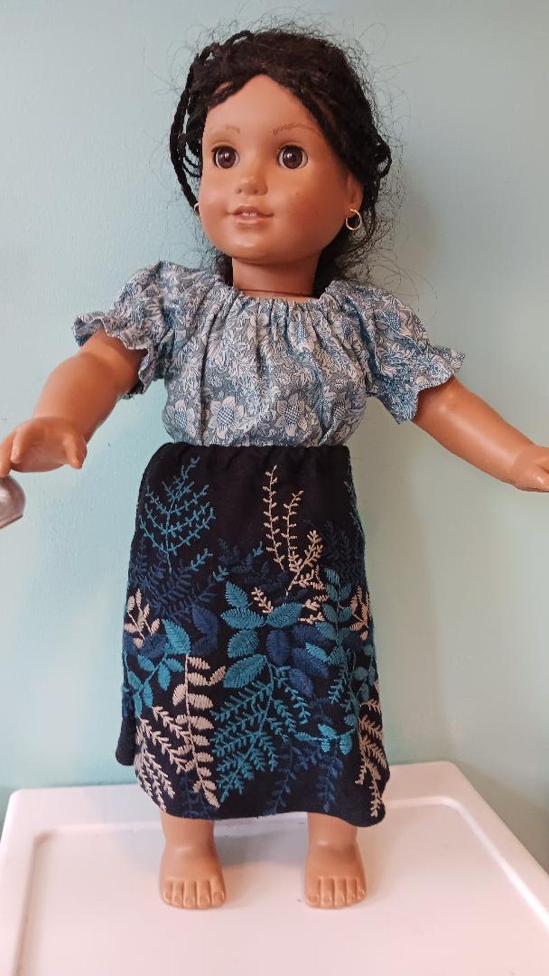 May include: A doll wearing a blue floral top and a black skirt with blue and white embroidered leaf designs. The doll has dark hair and brown eyes, and is wearing gold hoop earrings. The doll is standing on a white surface.