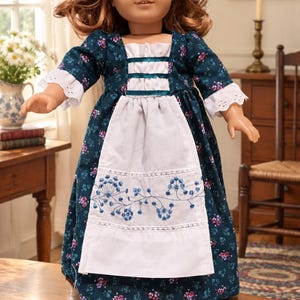 May include: A doll with wavy brown hair wearing a dark teal dress featuring a small floral pattern, white lace-trimmed sleeves, and a white apron with blue floral embroidery and horizontal teal ribbon accents on the bodice.