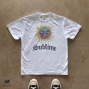 May include: A white short-sleeve t-shirt featuring a graphic of a colorful sun with a face and the word Sublime printed in black gothic-style lettering below the sun. The shirt has a crew neckline and is displayed with black and white sneakers.
