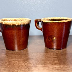 May include: Two ceramic mugs with a glossy dark brown finish and a cream-colored drip glaze along the upper rim. Each mug features a sturdy, angular handle attached to the side, with a tapered body that narrows toward the base.