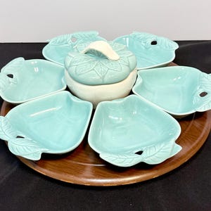 May include: A set of five light blue ceramic dishes shaped like apples, arranged around a central lidded bowl with a matching leaf-patterned lid. The entire collection sits on a circular wooden lazy susan tray with a smooth, dark brown finish.