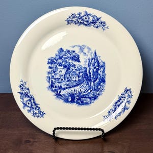 May include: A round cream-colored ceramic plate featuring a central blue transferware illustration of a rural landscape with houses and trees. The rim is decorated with three distinct blue floral clusters positioned at the top, bottom left, and bottom right.