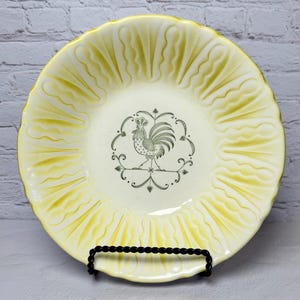 May include: A round ceramic bowl with a cream-colored center and a yellow scalloped rim featuring embossed vertical ridges. The center of the bowl displays a black line drawing of a rooster surrounded by a decorative circular frame of dots and leaves.