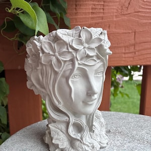 May include: A white concrete planter shaped like a human head, featuring detailed facial features and hair adorned with sculpted flowers and leaves. The vessel has an open top for plants and a textured surface, showcasing a monochromatic finish.