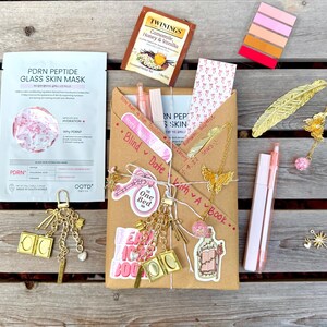 May include: A gift set featuring a PDRN Peptide Glass Skin mask, a Twinings Camomile Honey & Vanilla tea bag, a brown paper-wrapped book, gold-toned metal feather and butterfly bookmarks, pink pens, a book-shaped keychain, and colorful sticky note tabs.
