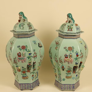 Pode incluir: Um par de potes de gengibre hexagonais de cerâmica na cor celadon com tampas. Cada tampa apresenta um pequeno leão esculpido. Os potes são decorados com motivos florais coloridos, borboletas e vários objetos em tons de rosa, azul e vermelho.