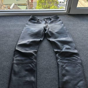 Puede incluir: Pantalones de cuero negro con un diseño clásico de cinco bolsillos, cierre de botones y detalles de costuras horizontales visibles en la zona del muslo. Los pantalones tienen un corte recto con un acabado mate liso y trabillas.