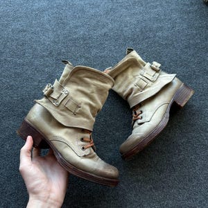 May include: A pair of beige leather ankle boots featuring a slouchy shaft, a decorative strap with a silver-toned metal buckle, and brown laces. The boots have a rounded toe, a low stacked heel, and a distressed finish on the leather surface.