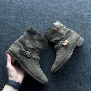 May include: A pair of olive green suede ankle boots featuring a rounded toe, a low stacked heel, and a decorative strap across the mid-foot. The strap is secured with a brass-toned metal buckle and accented with small, evenly spaced metal studs.