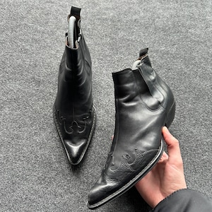 Può includere: Un paio di stivaletti in pelle nera con punta affusolata, tacco a blocco e cuciture decorative sulla tomaia. Gli stivaletti presentano una linguetta sul retro del colletto e una finitura liscia su tutta la struttura superiore.