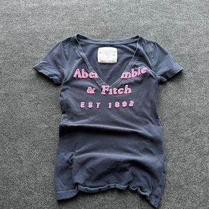 May include: A dark grey short-sleeve t-shirt with a V-neckline. The shirt features pink embroidered lettering that reads Abercrombie & Fitch EST 1892 across the chest. A white brand label is stitched inside the neckline of the garment.