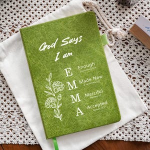 May include: A green journal with a textured cover featuring the white text God Says I am EMMA, with E for Enough, M for Made New, M for Merciful, and A for Accepted, accompanied by a white floral line drawing and a green ribbon bookmark.