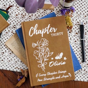 Puede incluir: Un diario de tapa dura marrón con texto blanco que dice Chapter THIRTY, el nombre Olivia y la frase Every Chapter Brings New Strength and Hope. La portada incluye una ilustración de línea blanca de flores y una mariposa.