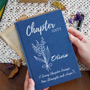Peut inclure: Un livre à couverture rigide bleu marine avec du texte blanc indiquant Chapter SIXTY, le prénom Olivia et la phrase Every Chapter Brings New Strength and Hope. Un dessin au trait blanc d'un muguet est centré sur la couverture avant.